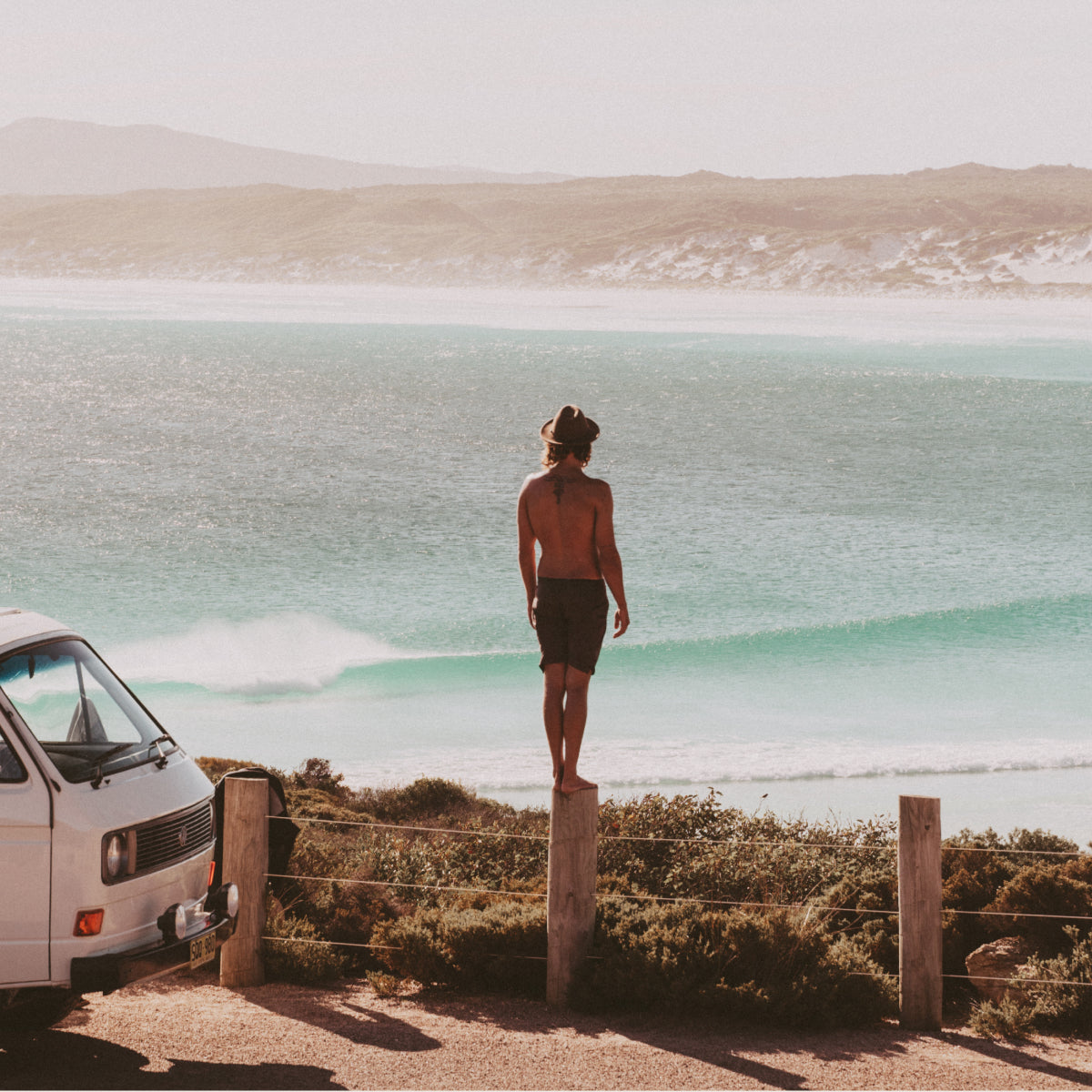 A man stands on top of a fence bollard next to his parked VW van, looking out at the ocean view