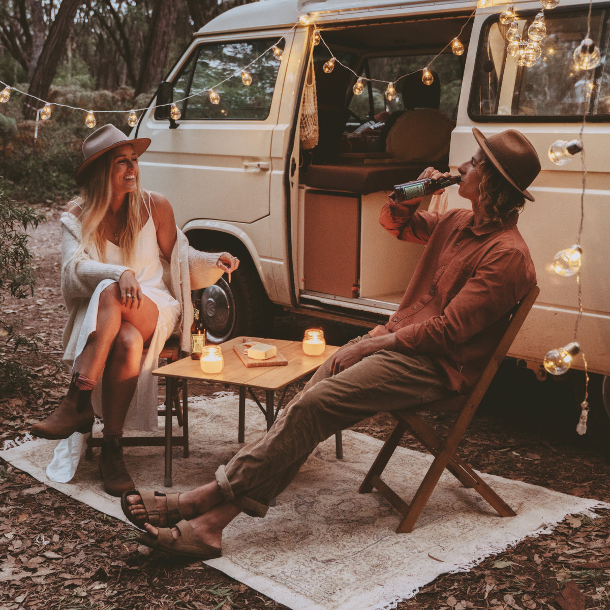 A couple sit next to their van drinking beer and eating cheese on outdoor chairs