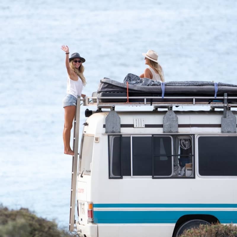 Two women climb on top of their camper van, one of them is waving to the camera and smiling