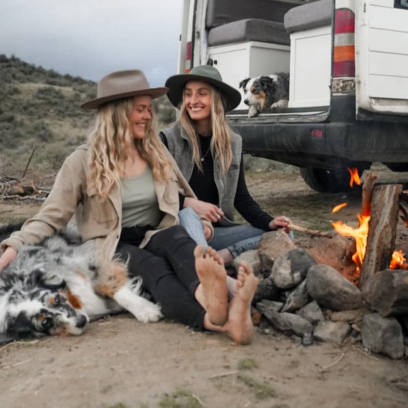 Two women sit with their dogs next to a camp fire they have set up behind their camper van