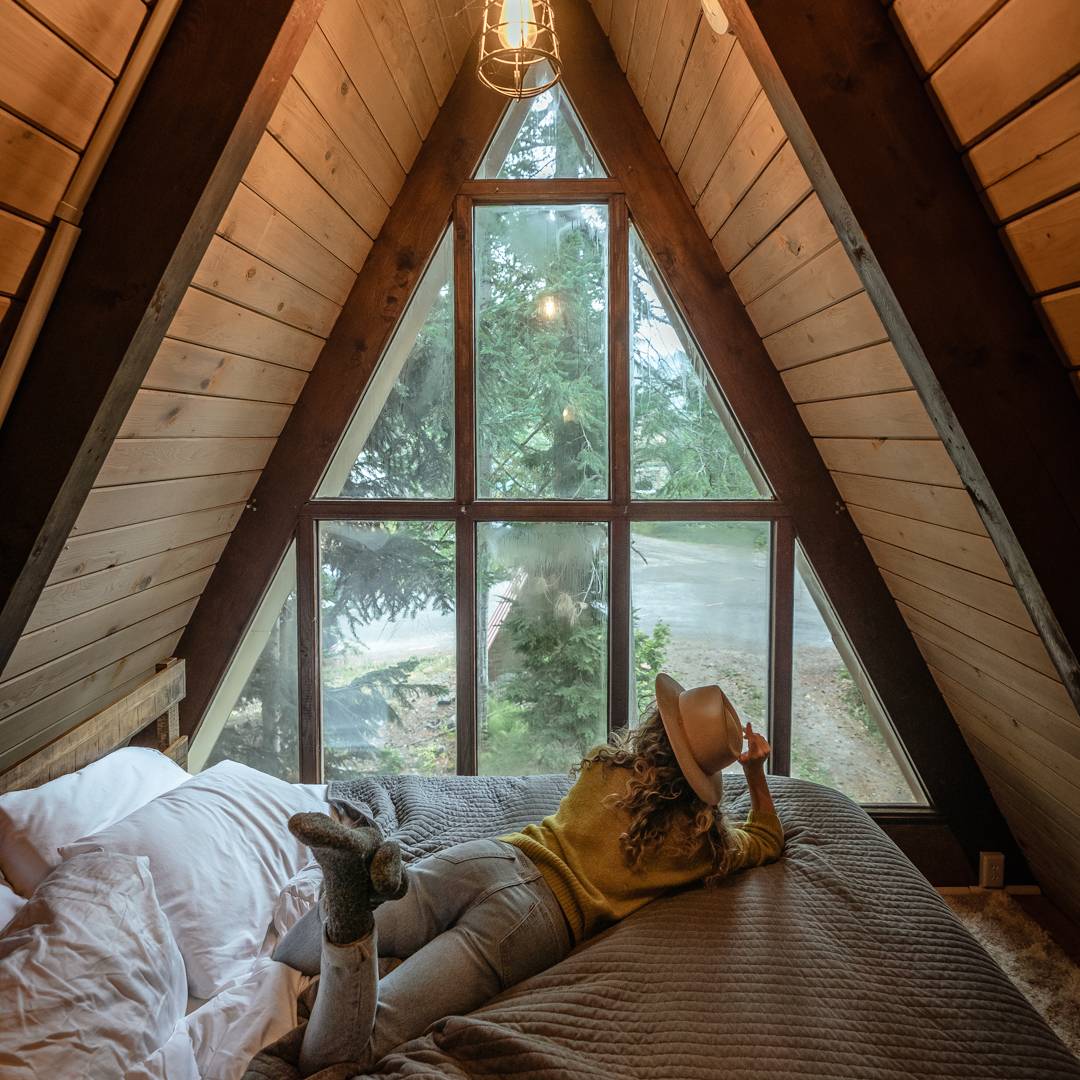Chelsea lies on the bed inside an A-frame cabin looking out the window