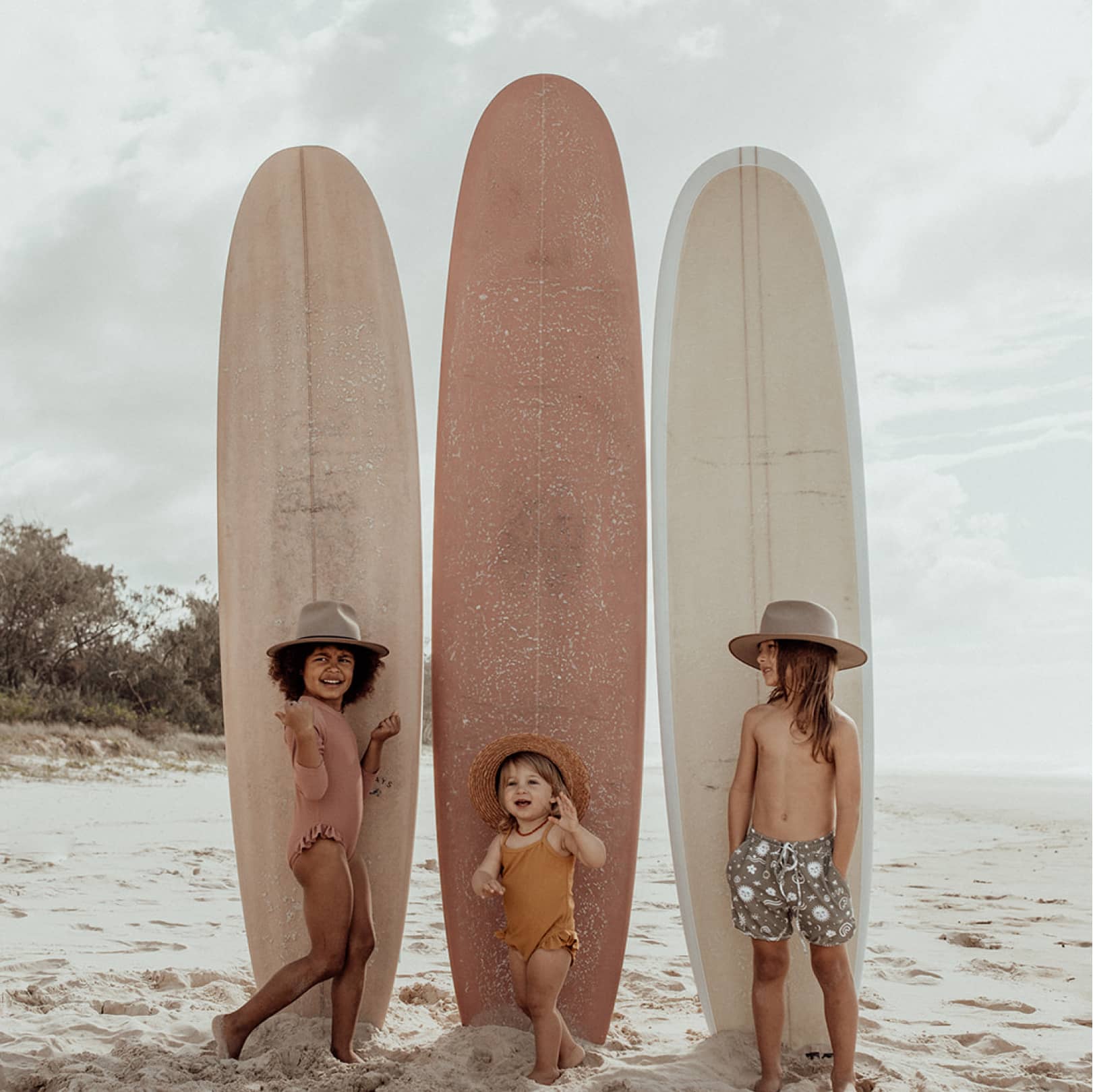 Three children wearing hats standing in front of surf boards on the beach