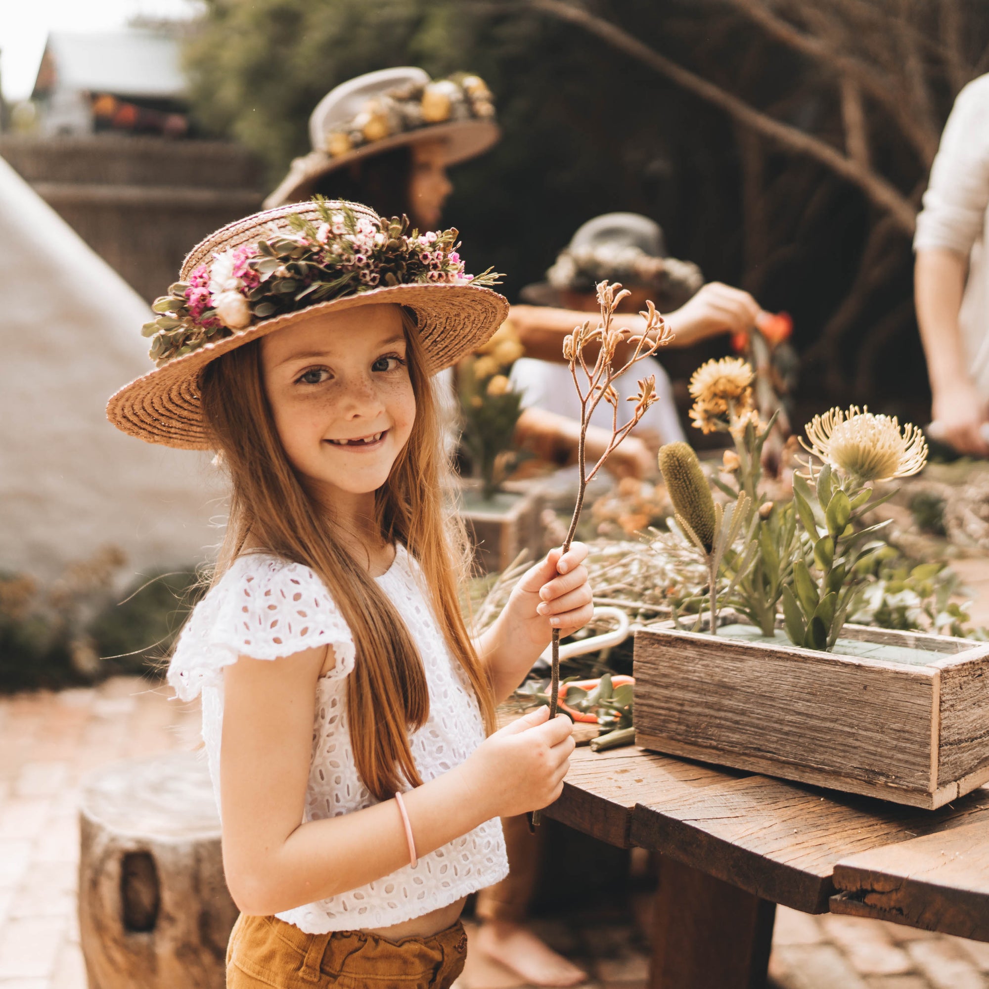A young girl wearing a wide brim straw hat is looking at the camera and holding up a flower stem with buds on the end