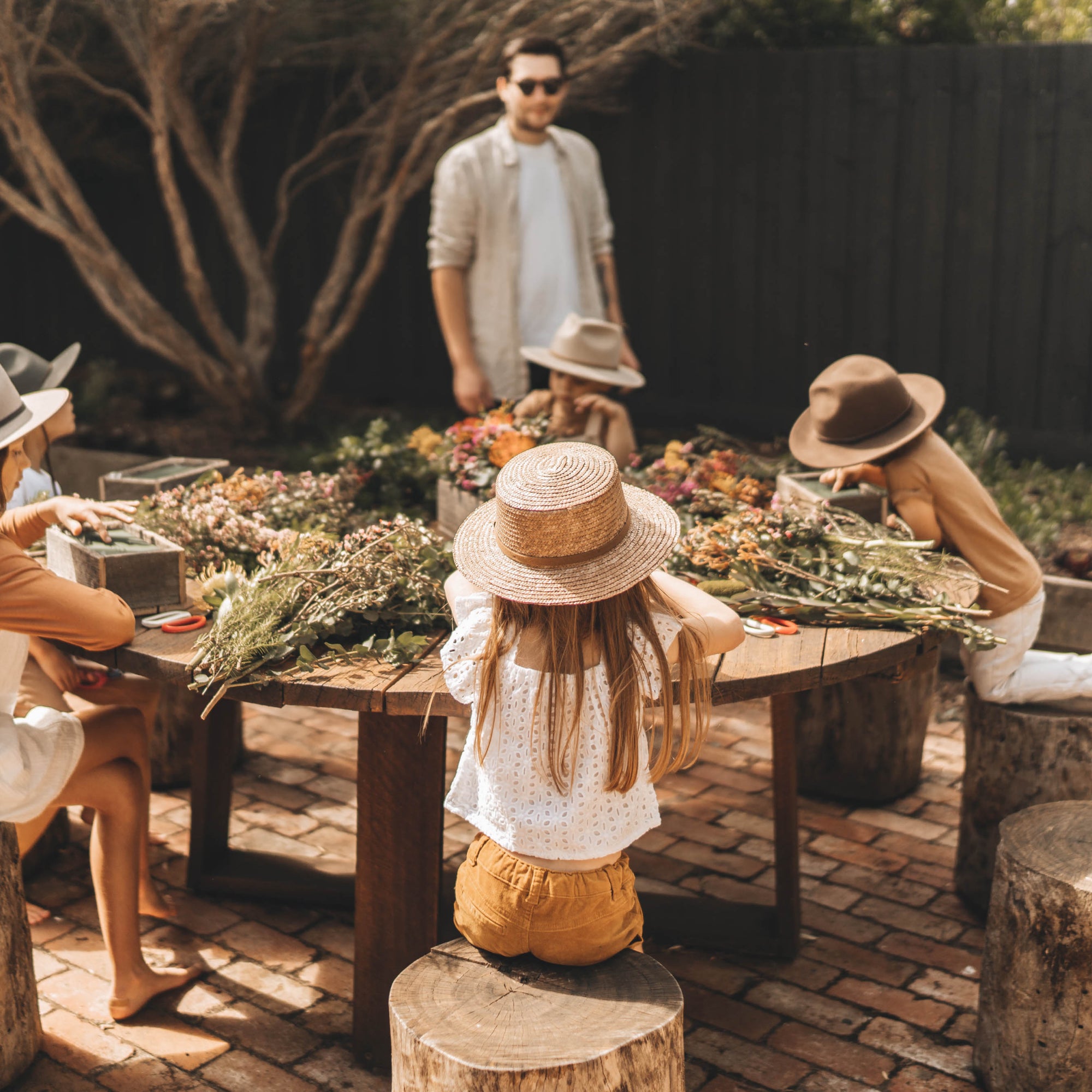 A group of young children sit around a large wooden table making flower arrangements for their wide brim wool hats