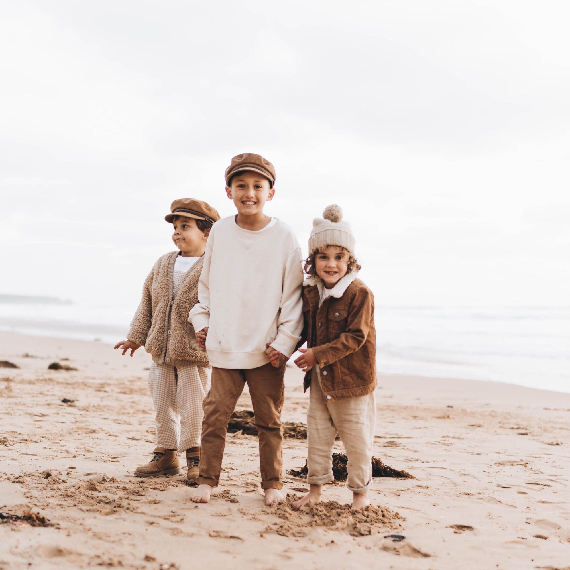 Three young boys are wearing hats and holding hands while standing on the beach