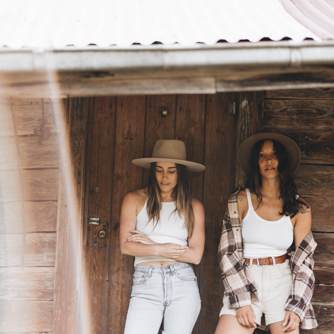 Two women standing in front of a cabin wearing wide brim wool hats