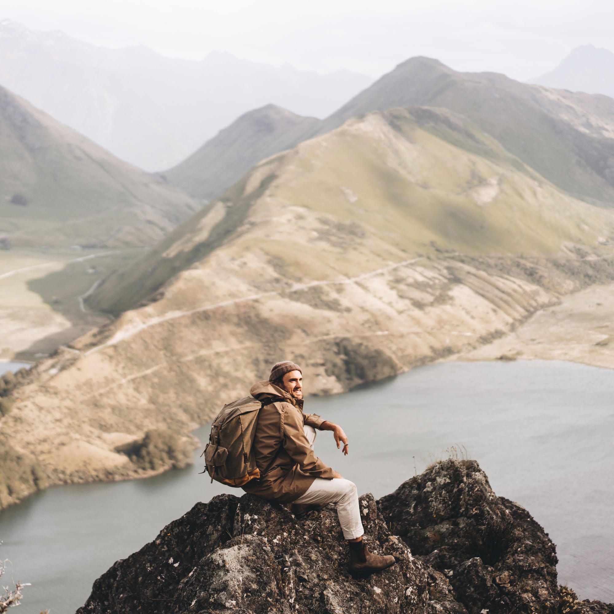 Alex wearing a backpack crouching down on the top of a mountain looking down at a view of the valley