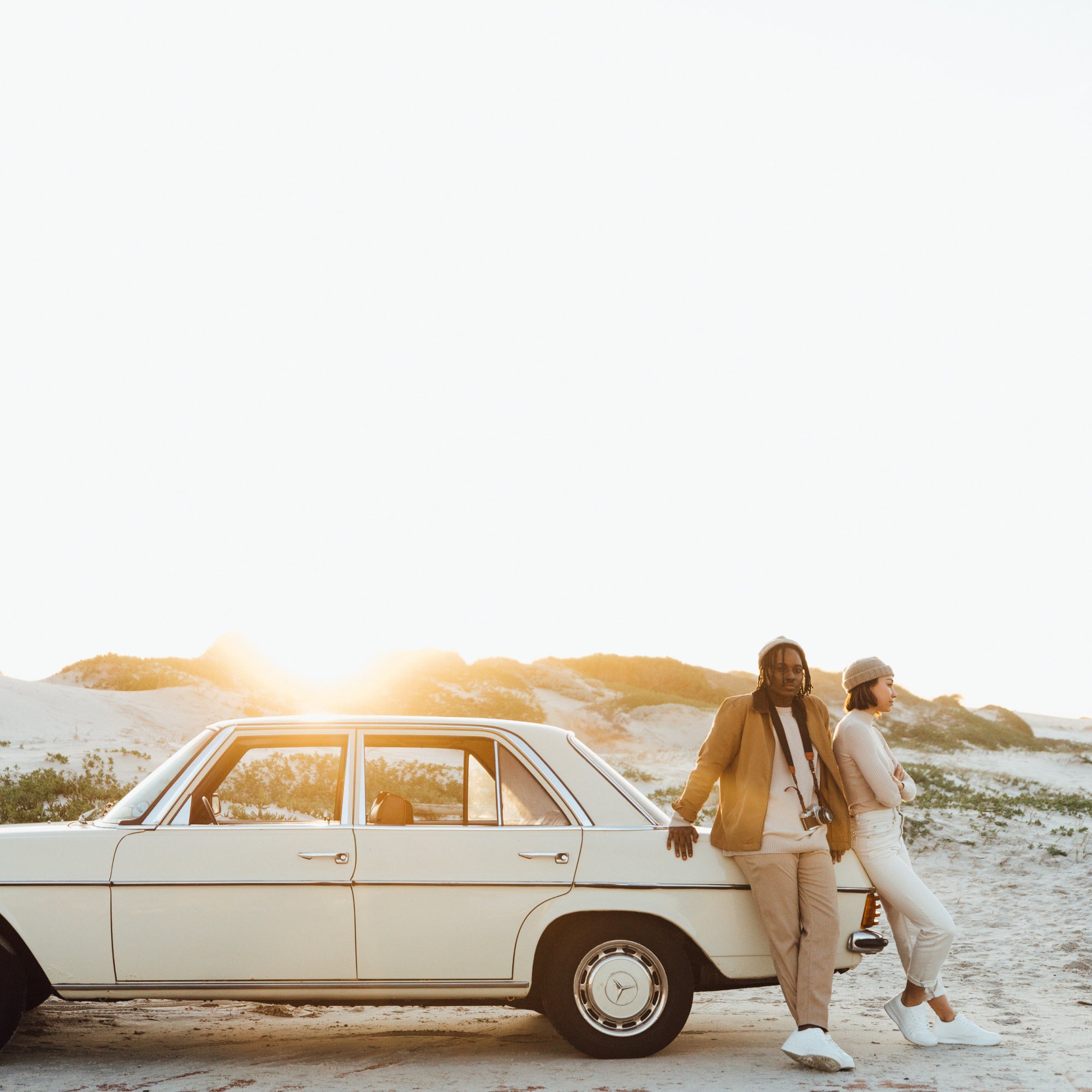 A man and a woman lean against the bonnet of a car parked on the beach, the man has a camera hanging around his neck