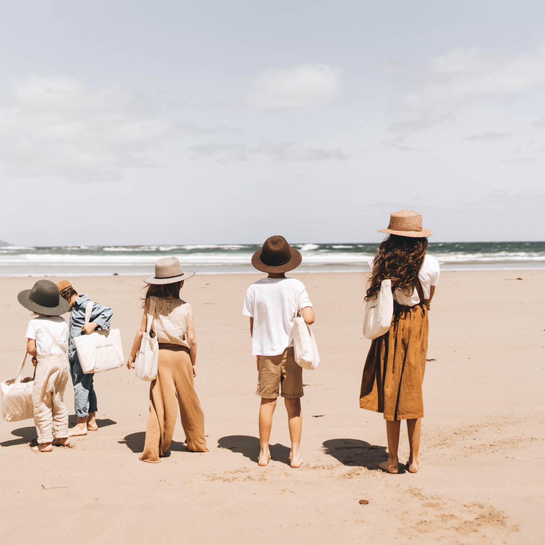 Five children are walking along the beach wearing wide brim hats and carrying tote bags