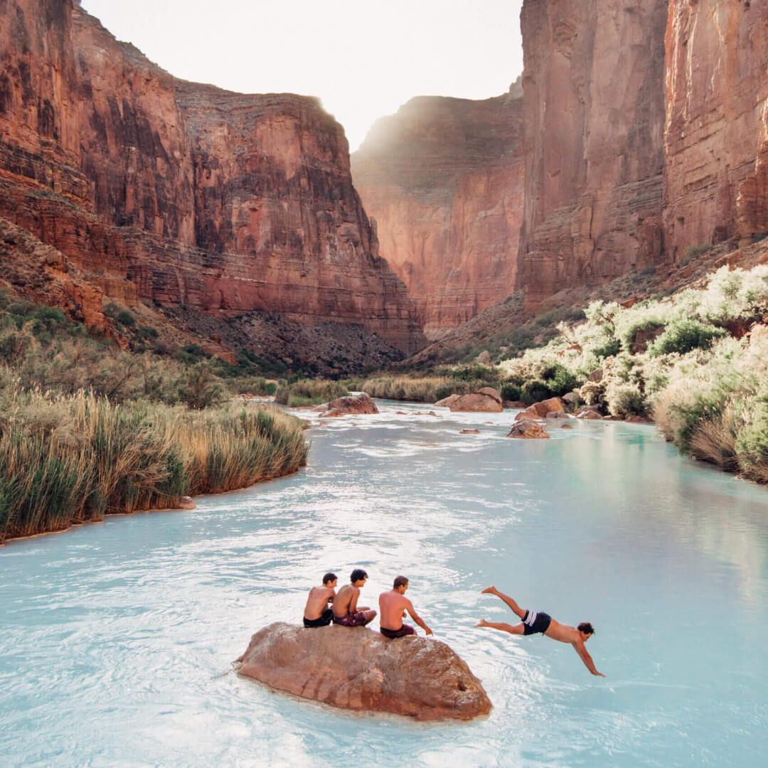 A group of people sitting on a rock in the middle of a gorge, one of them is diving into the water