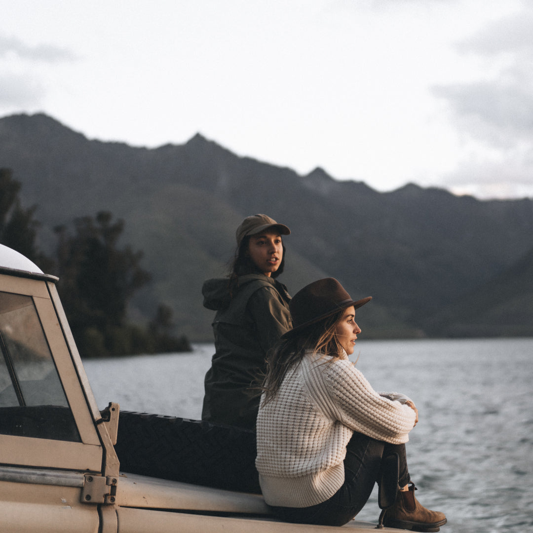 Two women sit on the bonnet of a parked Land Rover looking out at the views of the water in front of them