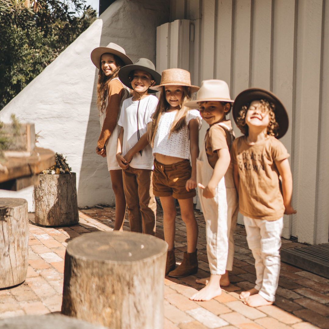 Five children line up in the sun next to a cabin all wearing wide brim hats