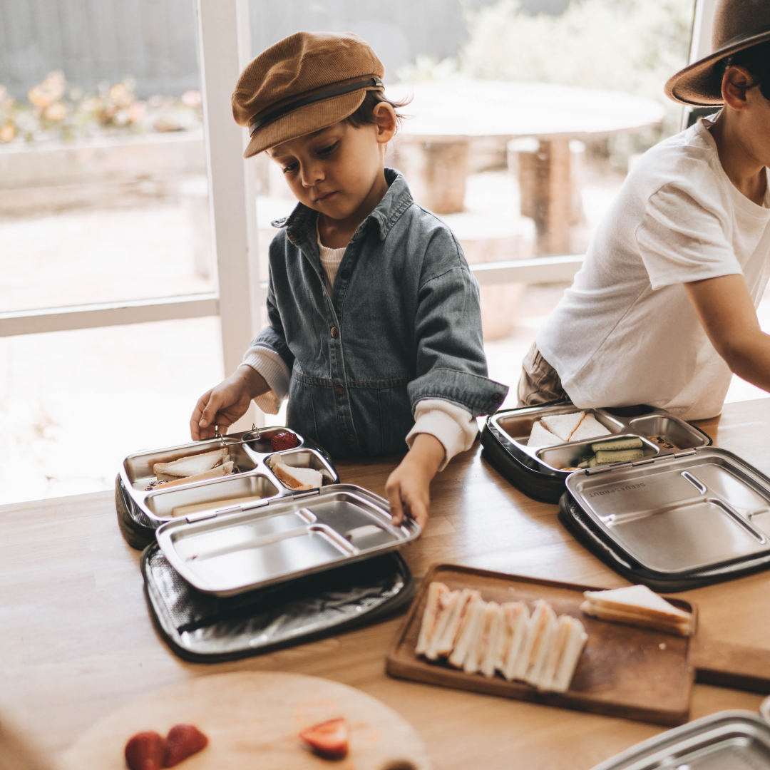 A young boy wearing a fisherman hat is packing food into his metal lunchbox