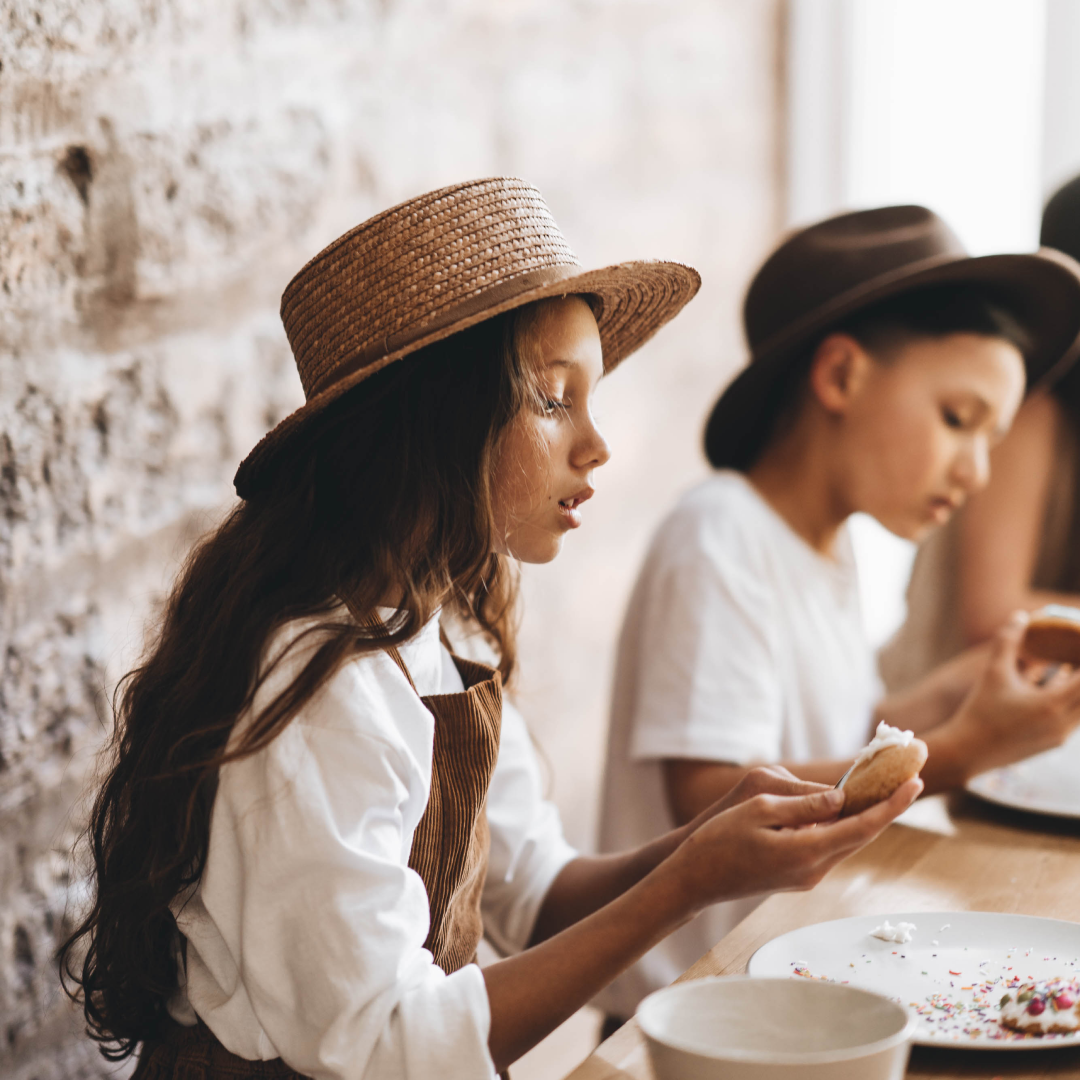 A young girl wearing a wide brim straw hat is putting icing on the inside of a cookie