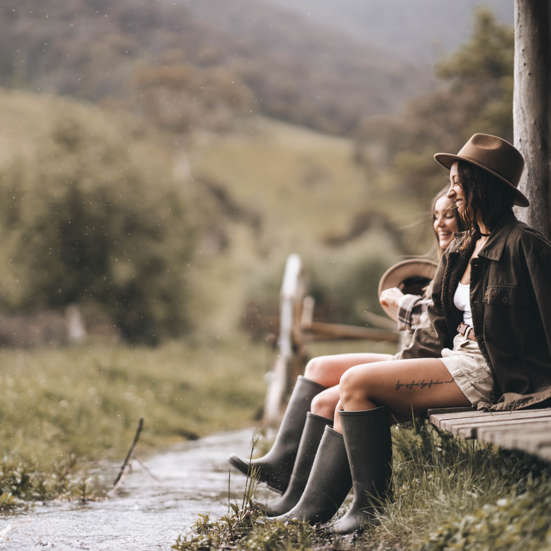 Two women sitting by a creek wearing wide brim wool hats and gum goots