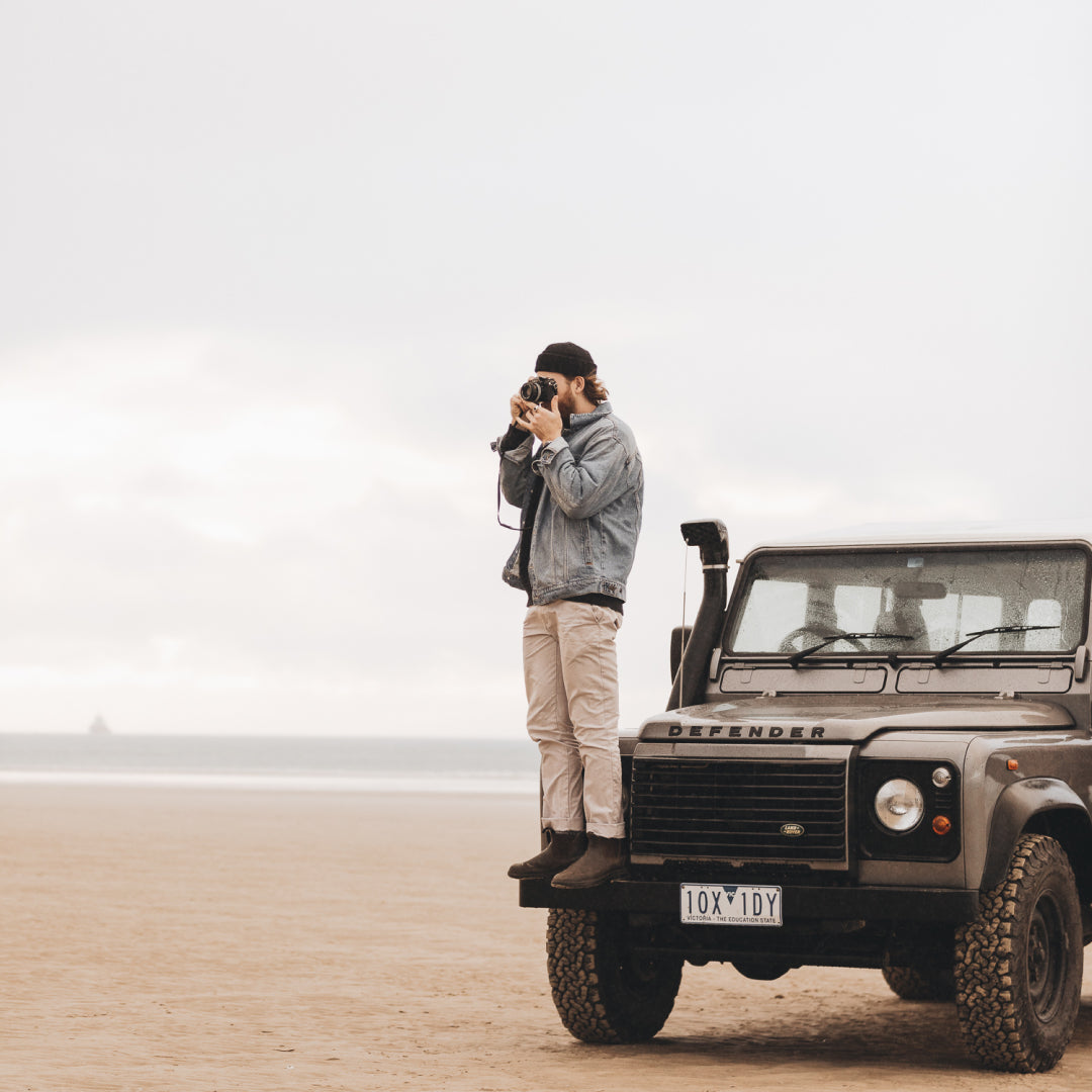 A man taking a photo standing on top of a land rover at the beach