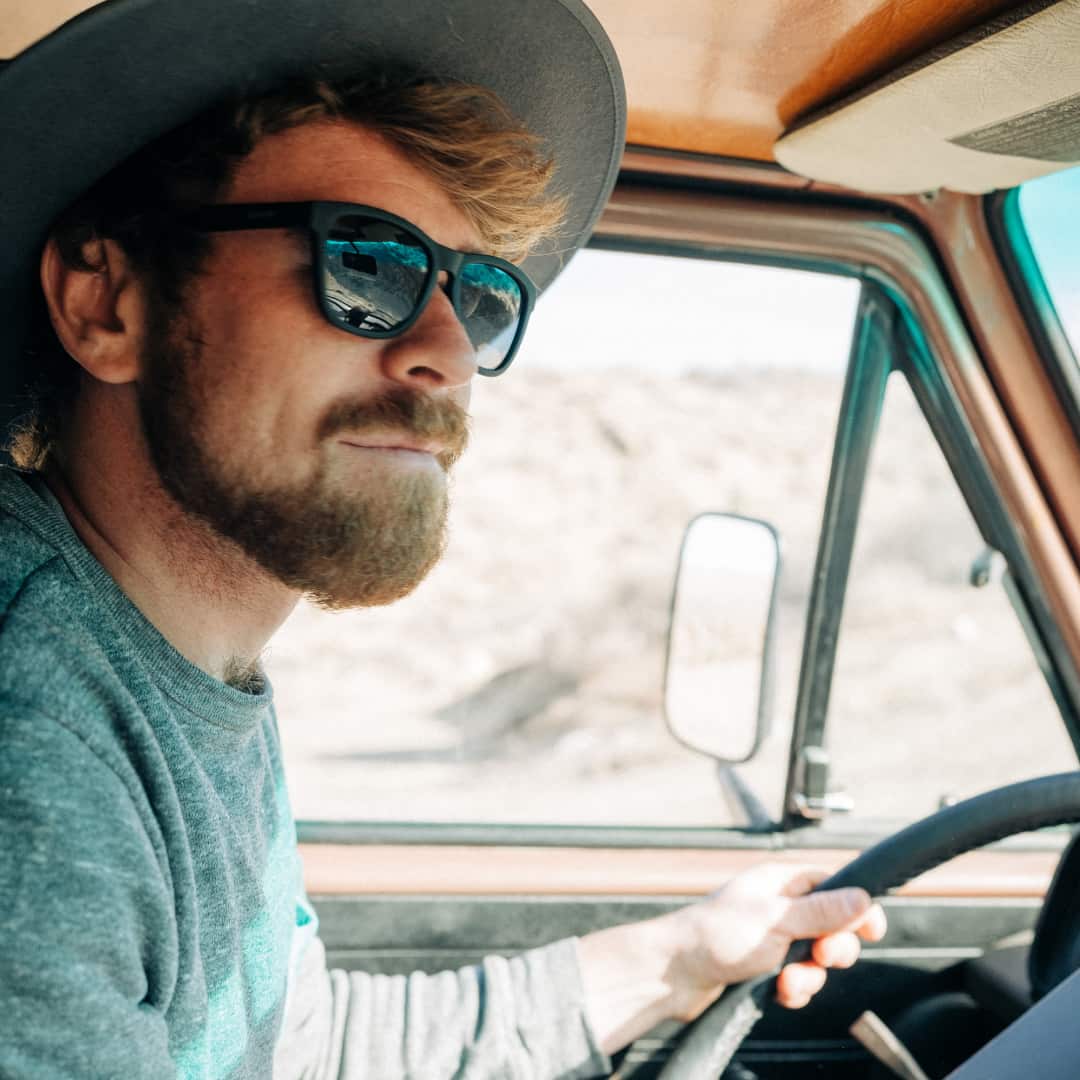 Close up photo of a man driving a truck through Joshua Tree wearing a wide brim hat and sunglasses