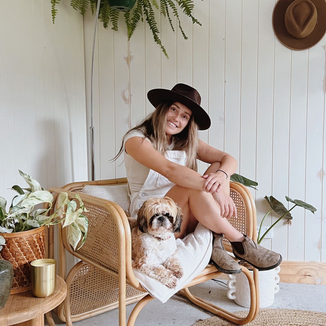 Woman with long brown and blonde hair wearing a wide brim wool hat sitting on a chair with her small fluffy dog