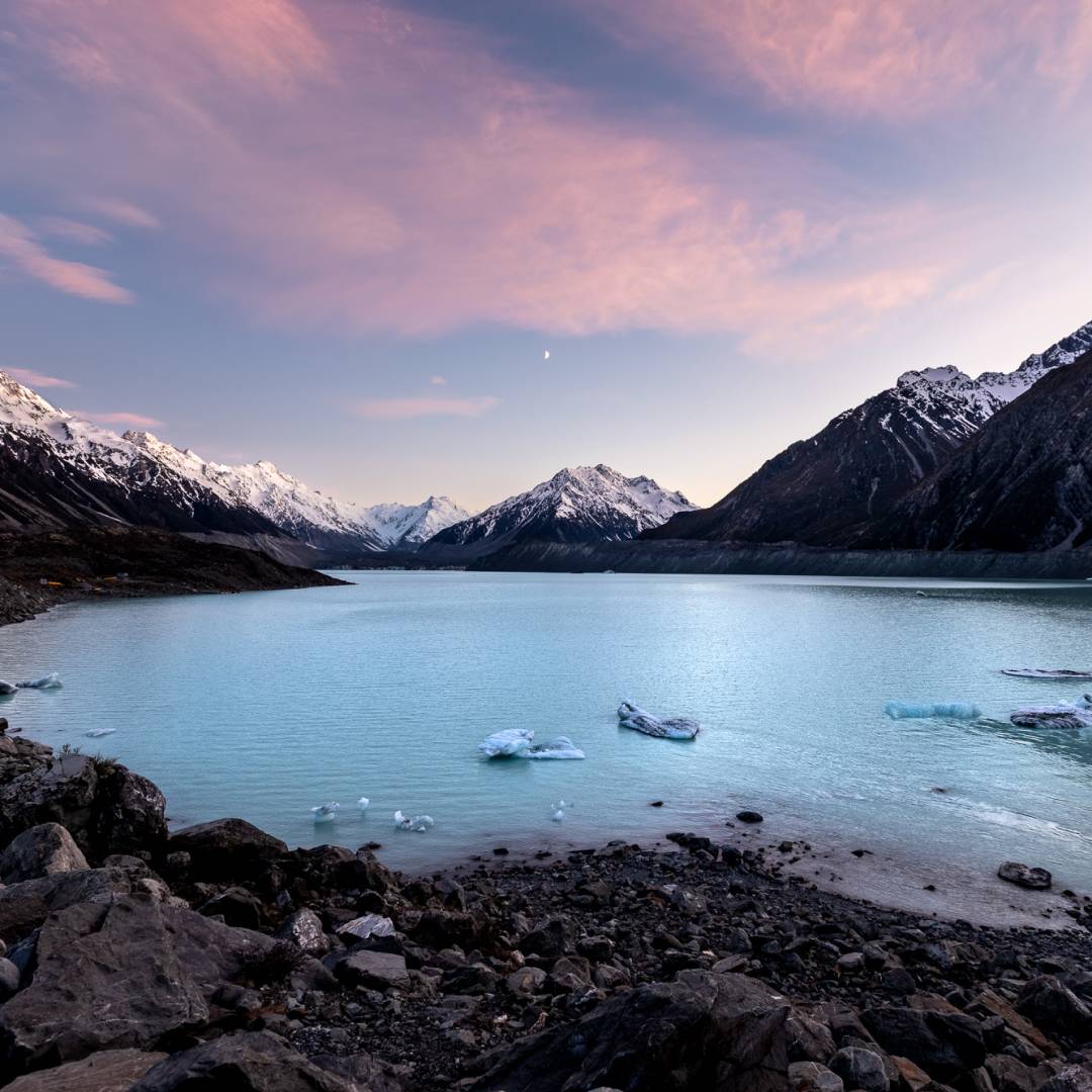 Rocky shore leading into a bright blue waterway at dusk, with snowcapped mountains in the background