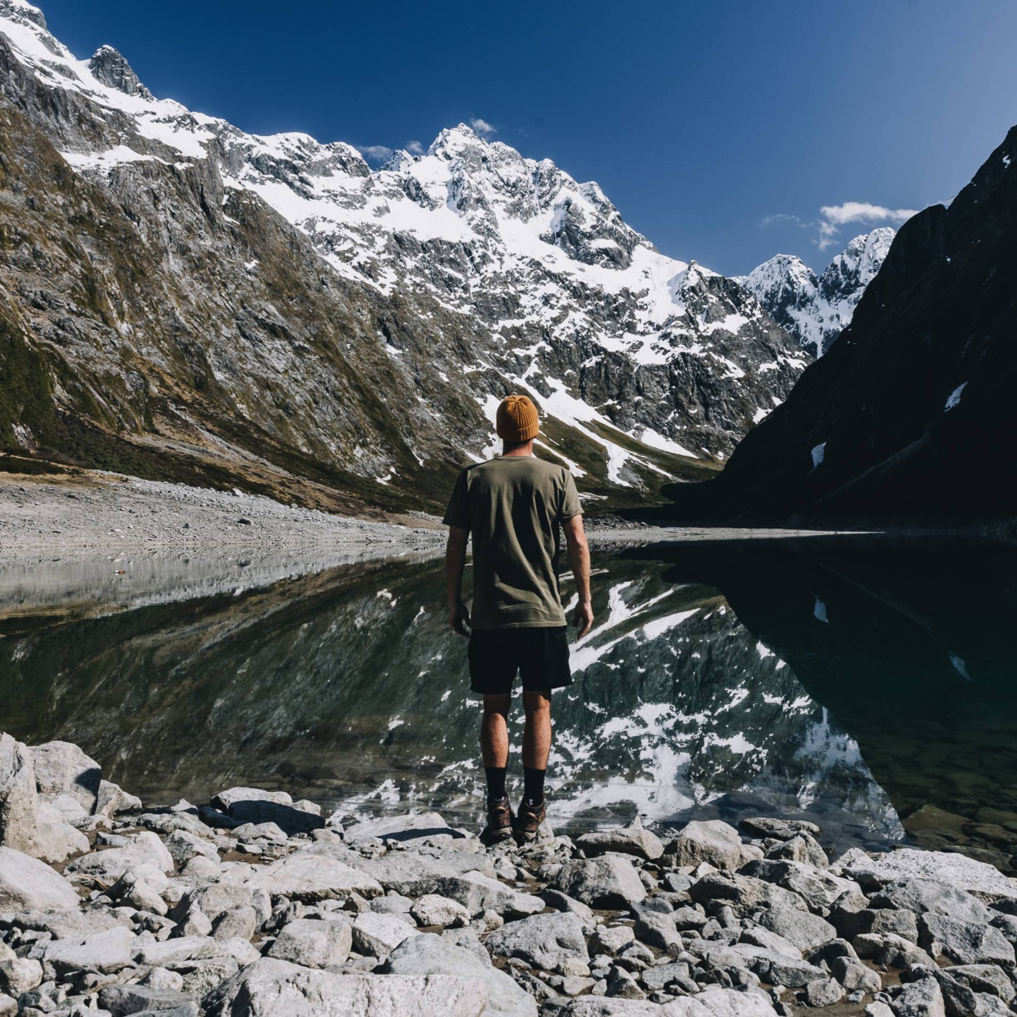 Marc is standing with his back turned to the camera looking out towards all the snow capped mountains behind the lake