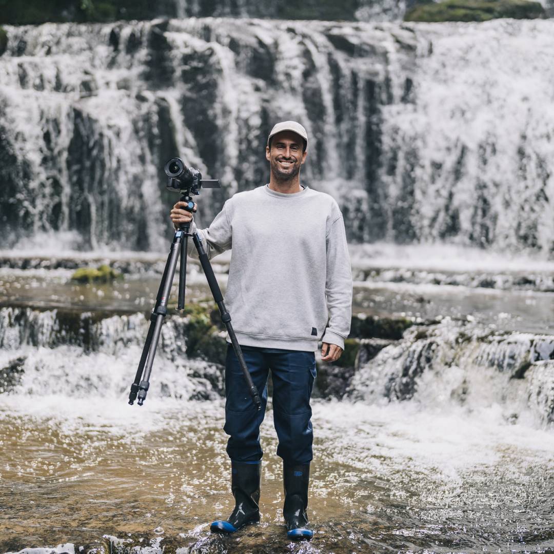 Marc holding up his camera and tripod standing in front of a waterfall