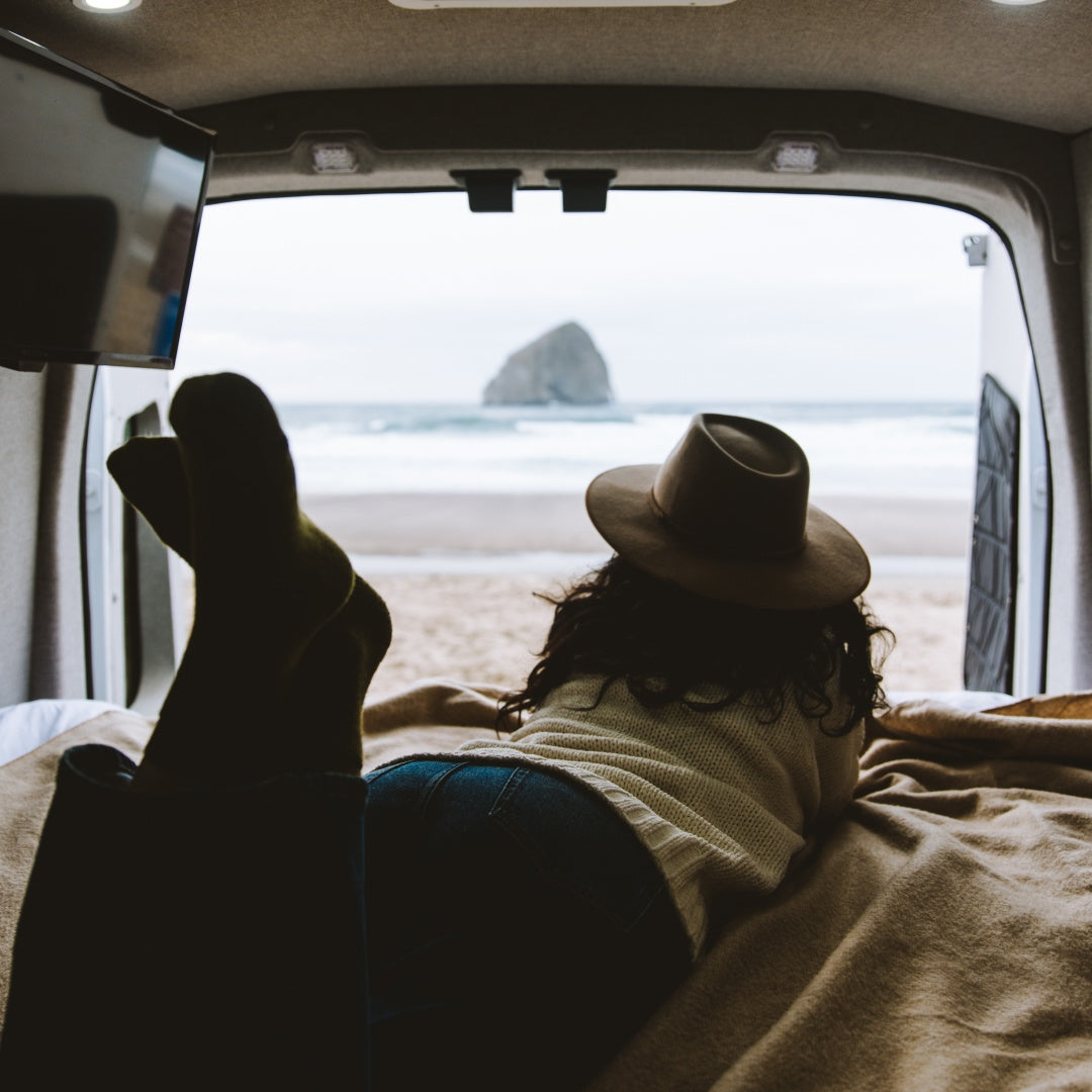 A woman is sitting inside the back of her campervan looking out at the beach