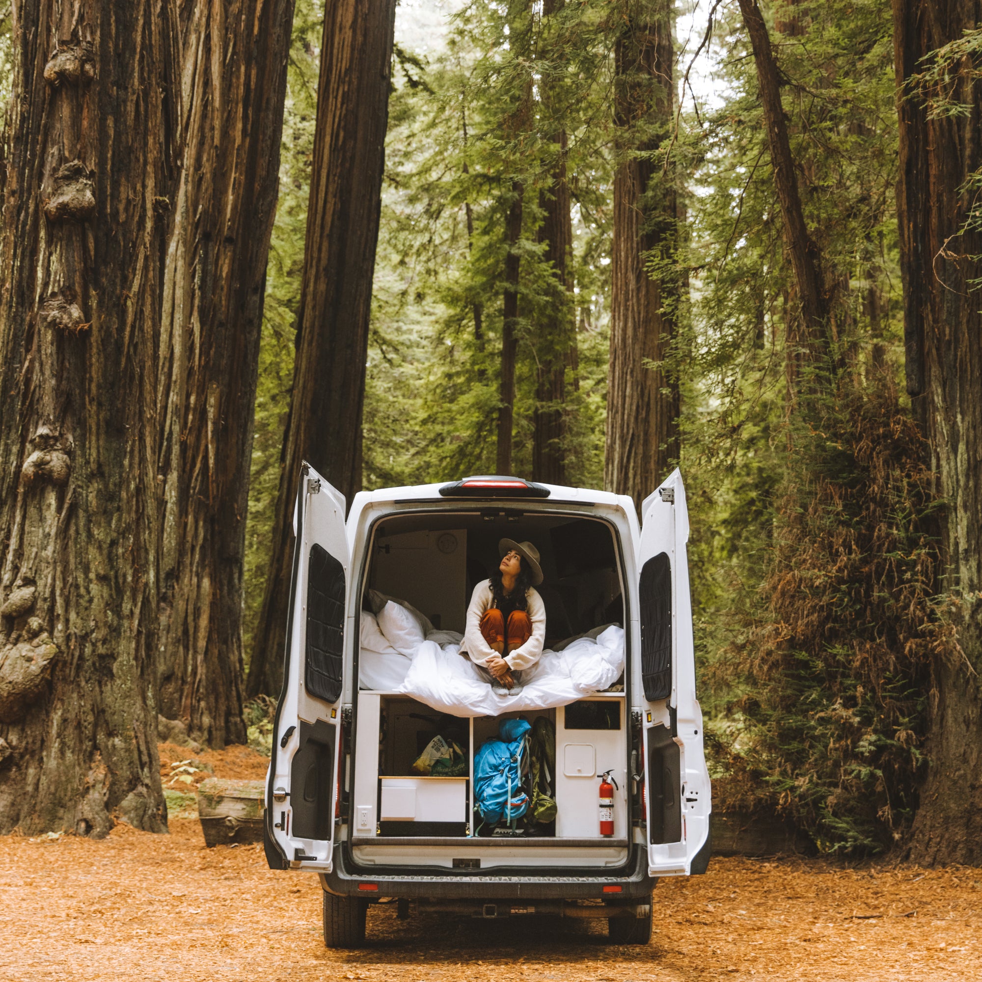 A woman wearing a wide brim wool hat is sitting in the back of her camper van parked amongst the redwood trees
