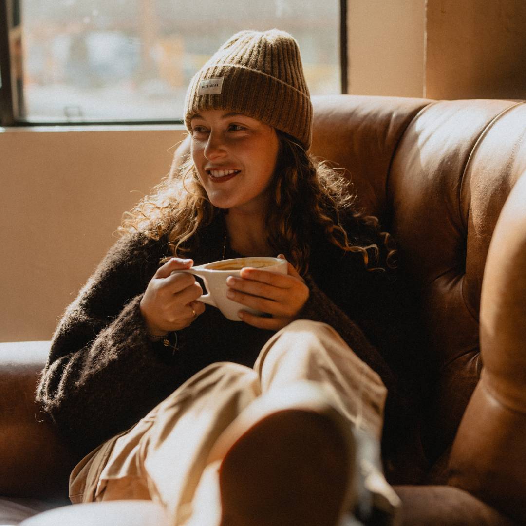 Woman with long, curly hair wearing a beanie and drinking a coffee