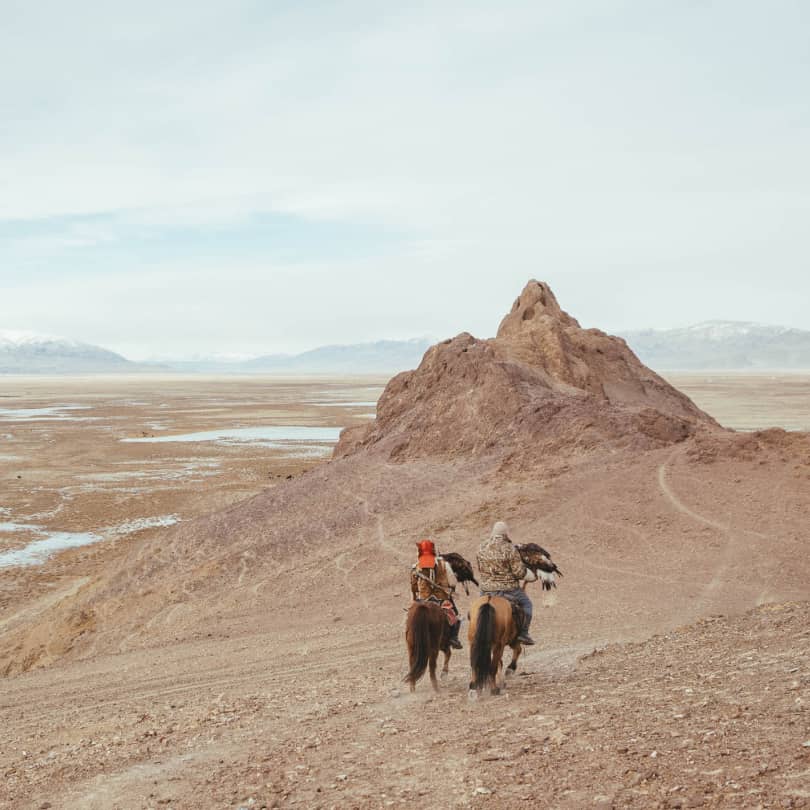 Mongolian men riding horses through mountains while holding eagles on their hands
