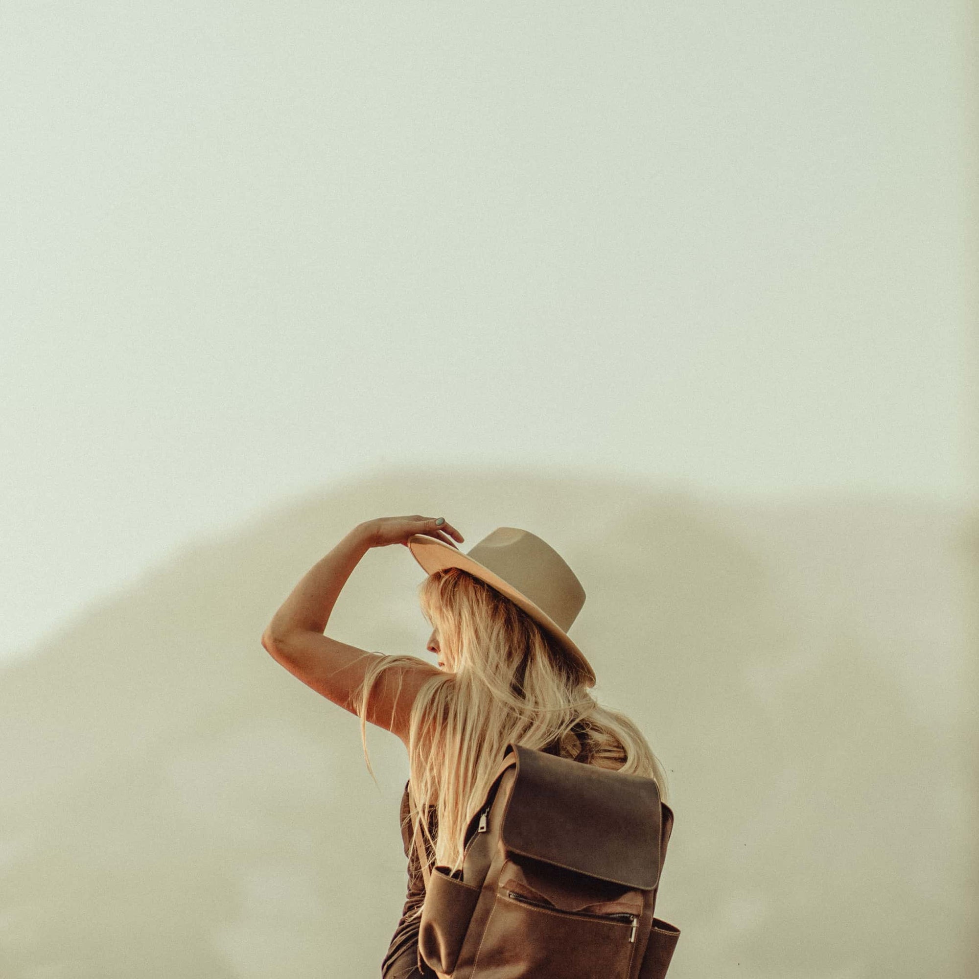 A woman wearing a backpack holds on to the front of her wide brim hat while hiking through a national park