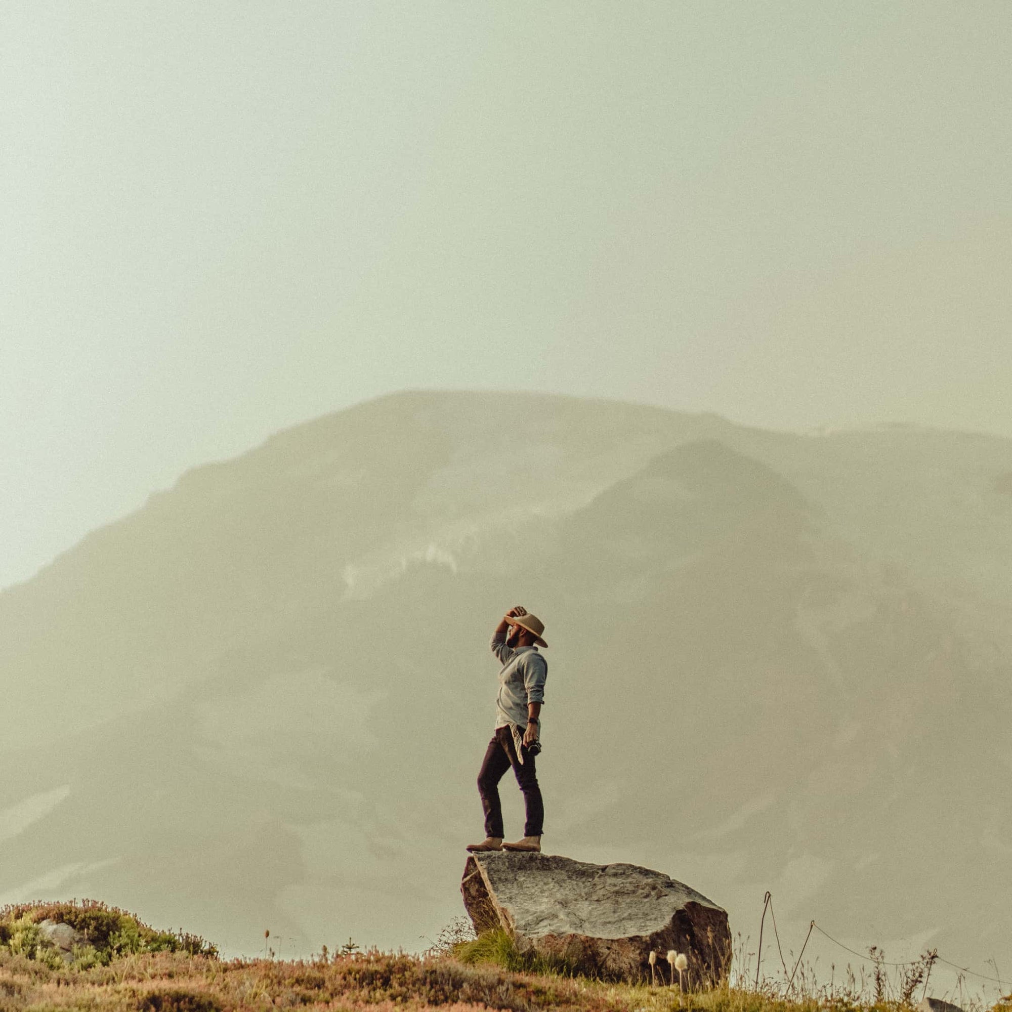 Drake standing on top of a rock holding his wide brim hat on top of his head