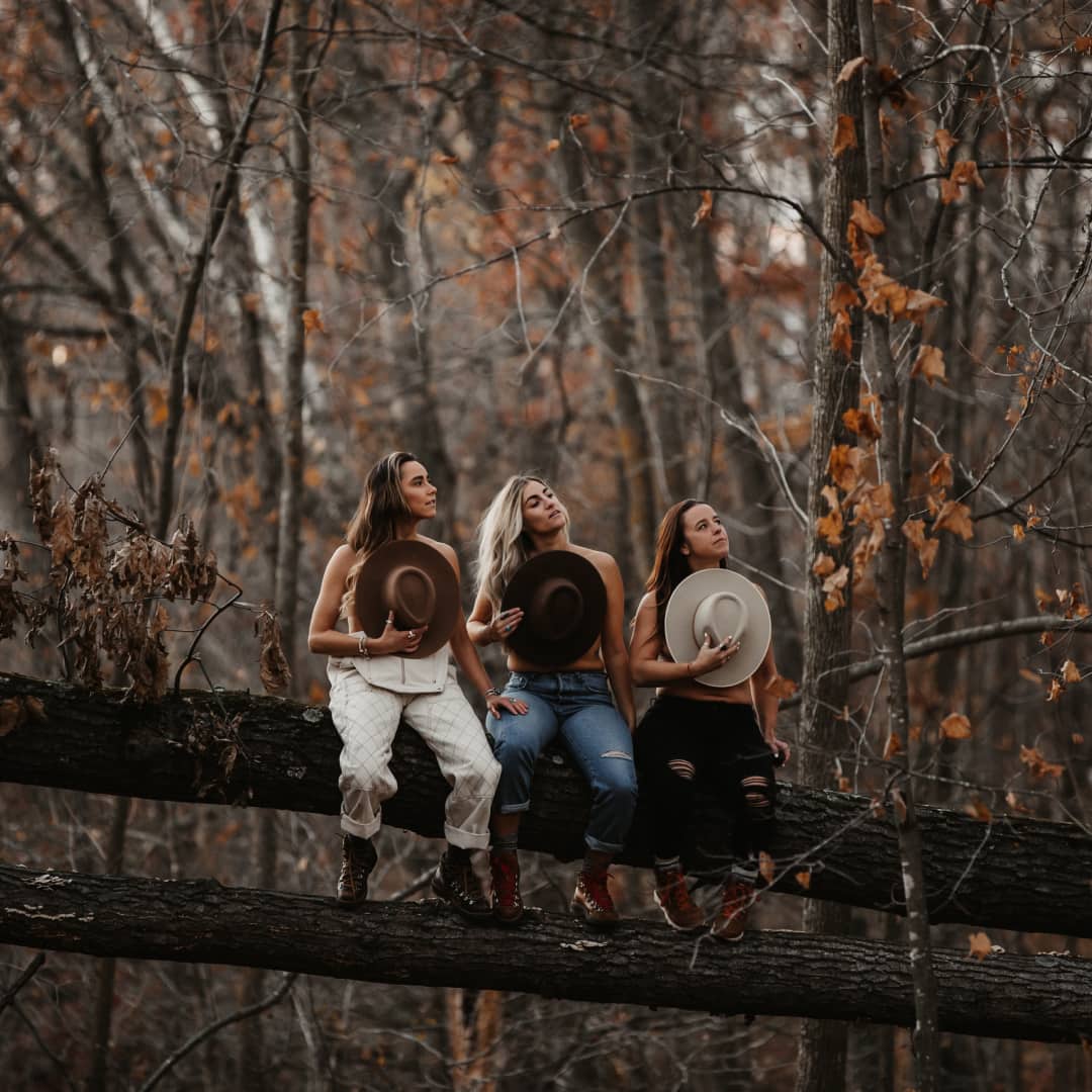Amanda, Taryn and Kaylee sitting together on a large fallen tree trunk, holding wide brim hats up to their chests