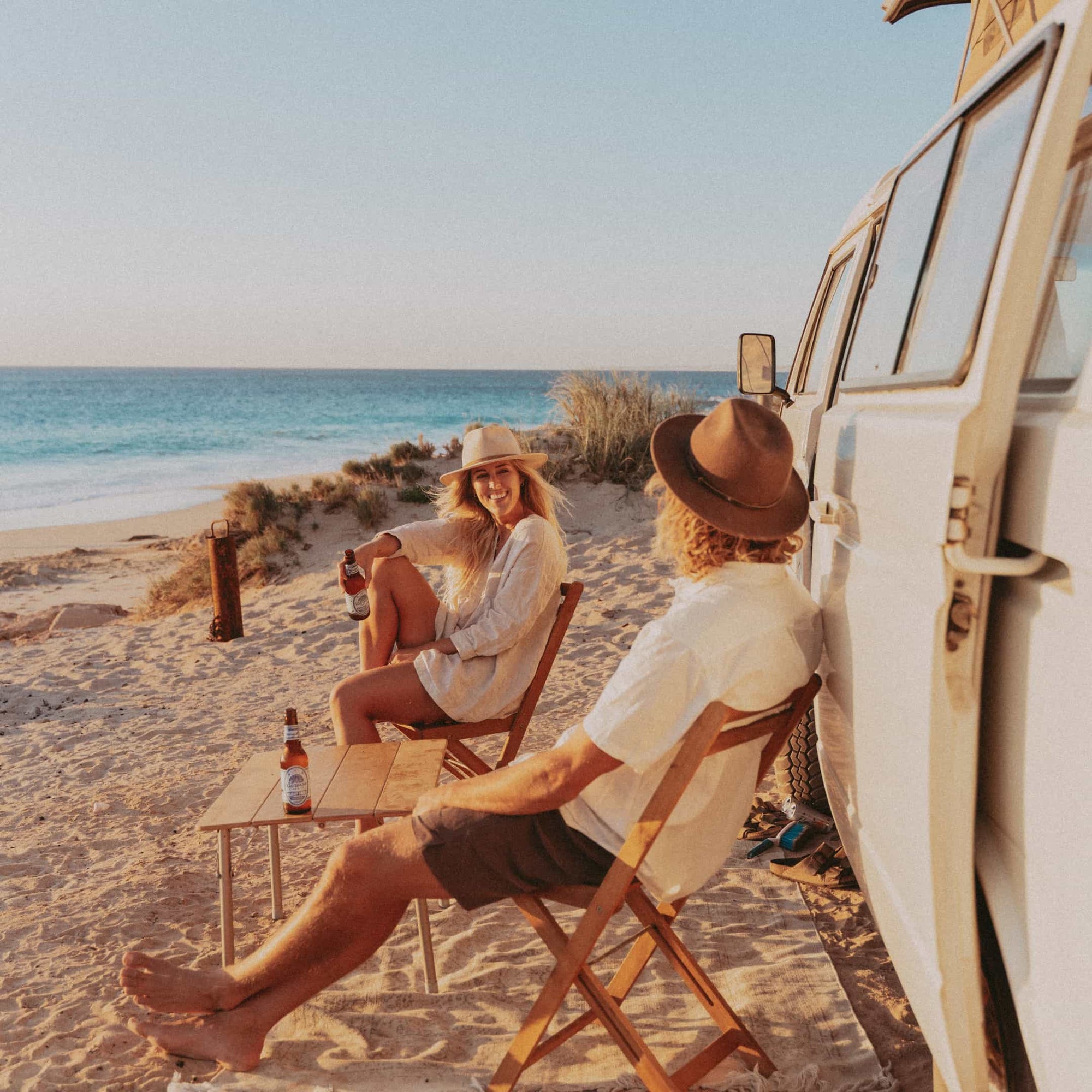 A young couple sit on the beach next to their VW can drinking beers