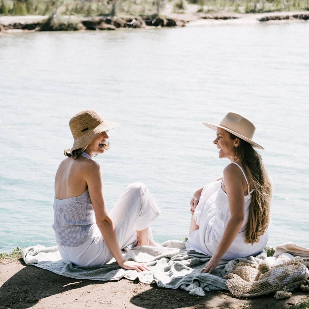 Two women sit on a picnic blanket at the beach smiling at each other