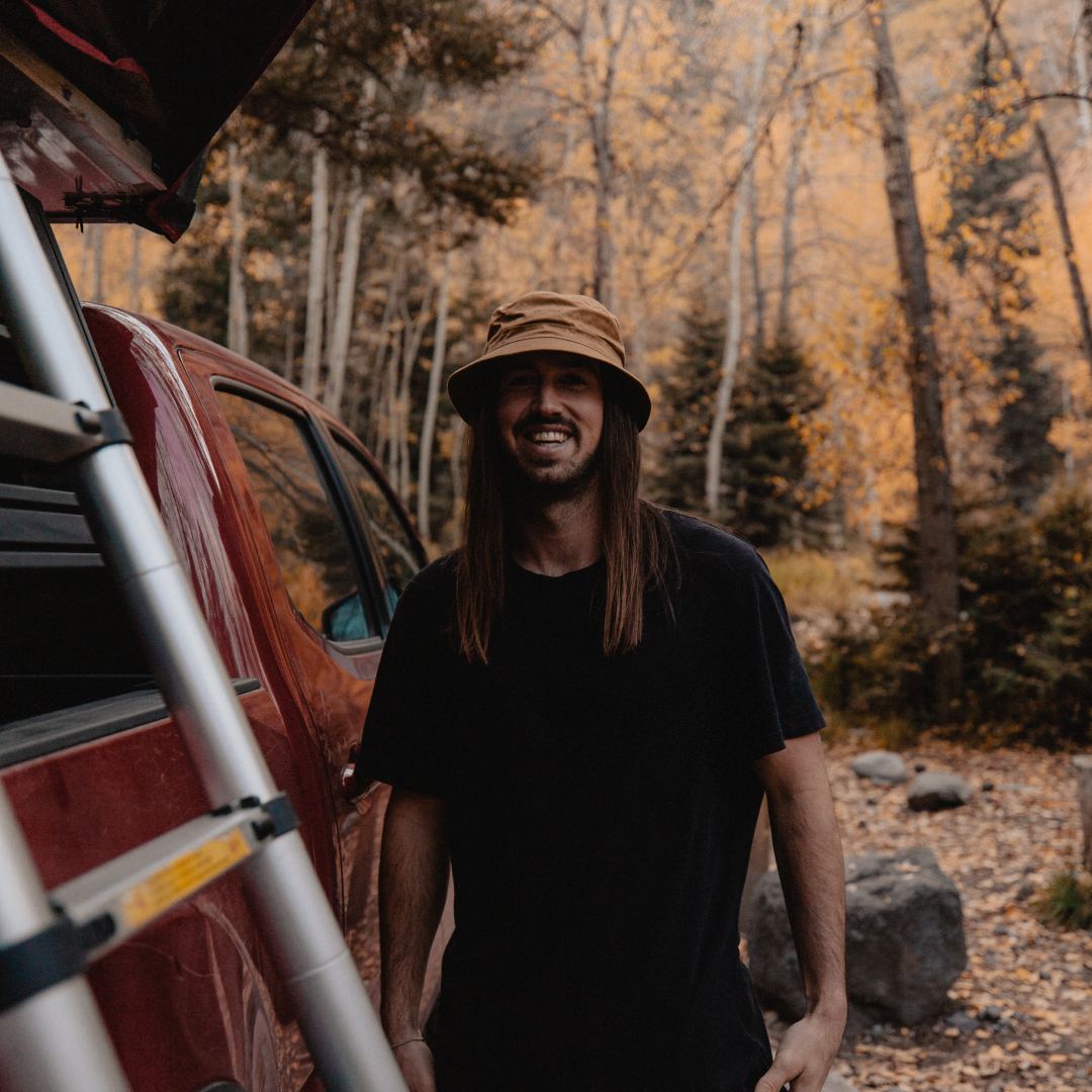 A man with long hair wearing a bucket hat smiling and standing next to a utility vehicle in the forest
