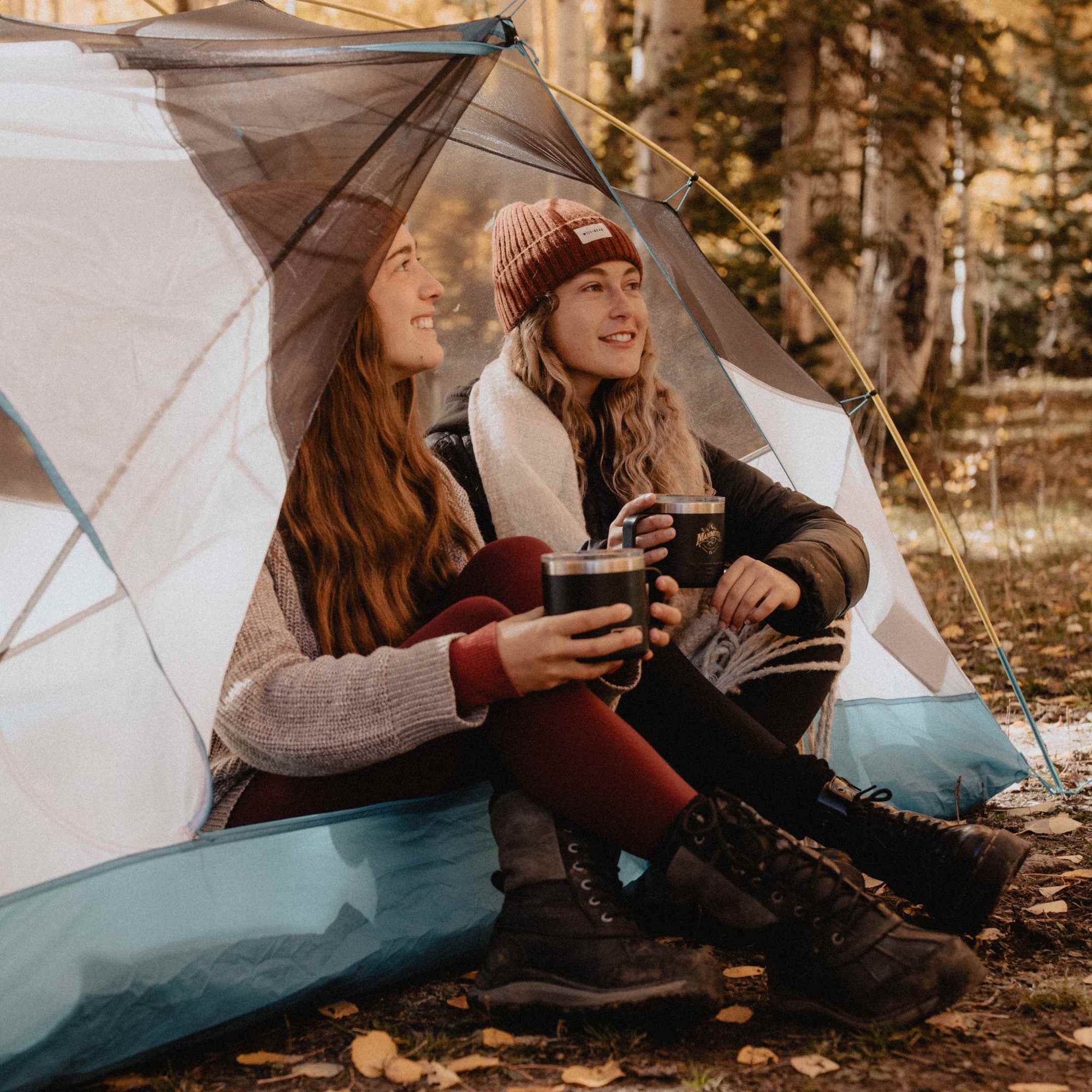 Two women wearing beanies sitting inside a tent drinking hot coffee