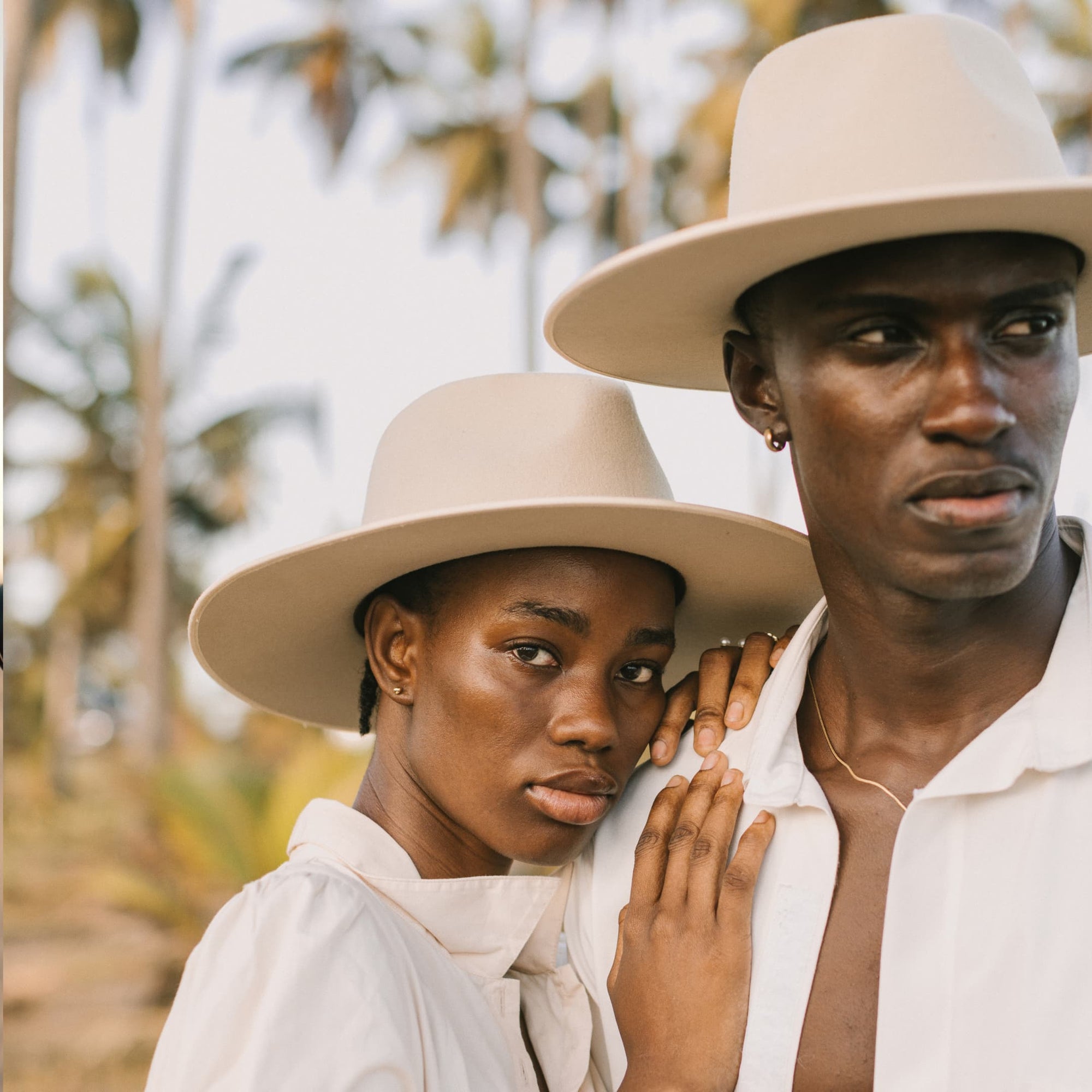 Close up photo of a man and woman wearing wide brim wool hats with palm trees behind them