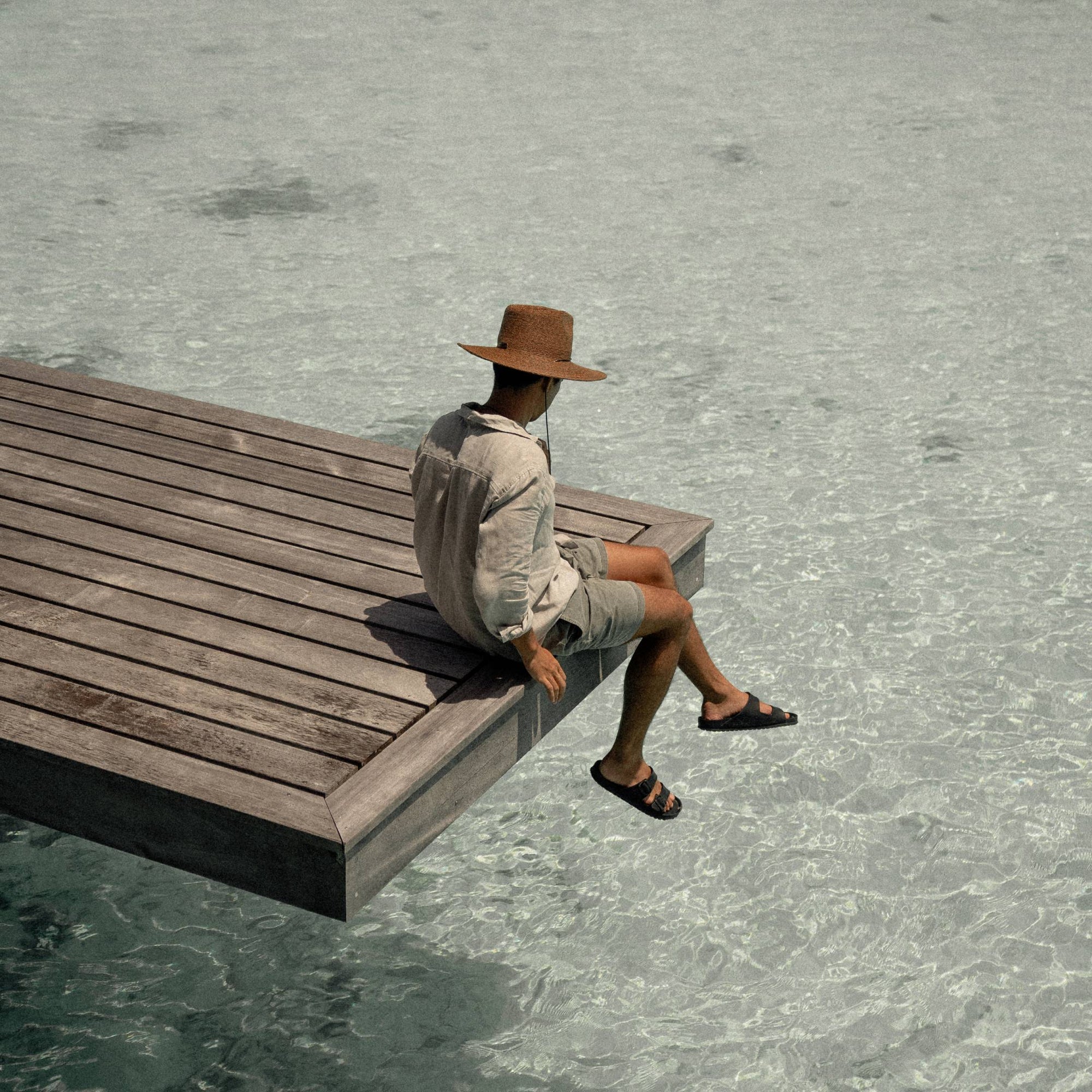 A man with short black hair wearing a wide brim straw hat, he is sitting on the edge of a jetty.