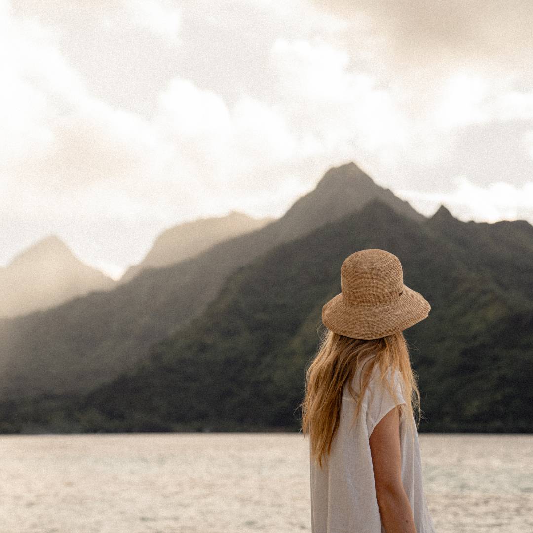 A woman with long blonde hair wearing a crocheted straw bucket hat is looking towards a mountain view.