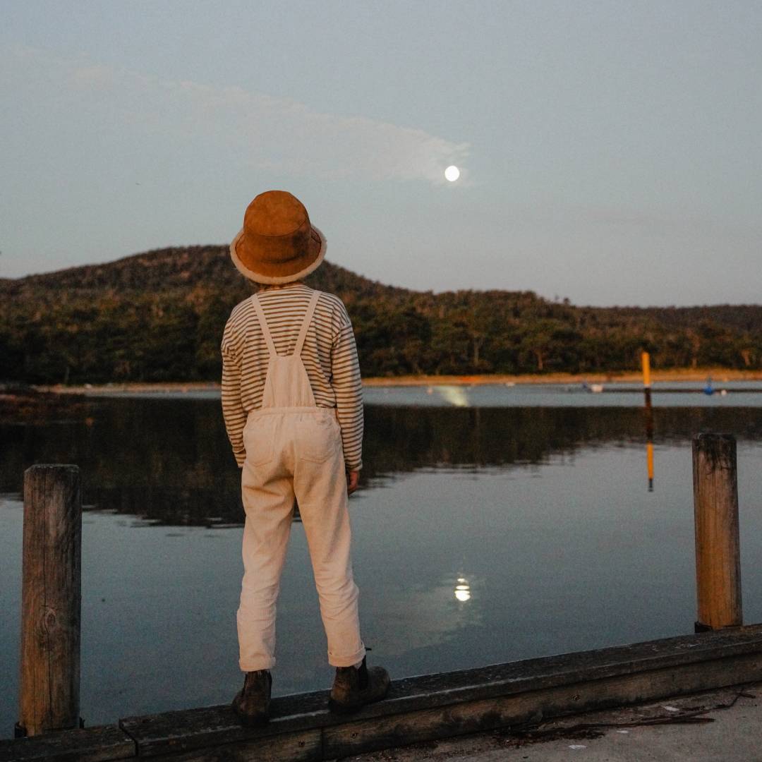 A young boy in overalls and an ugg wool bucket hat is standing on a pier looking at the fool moon behind the water
