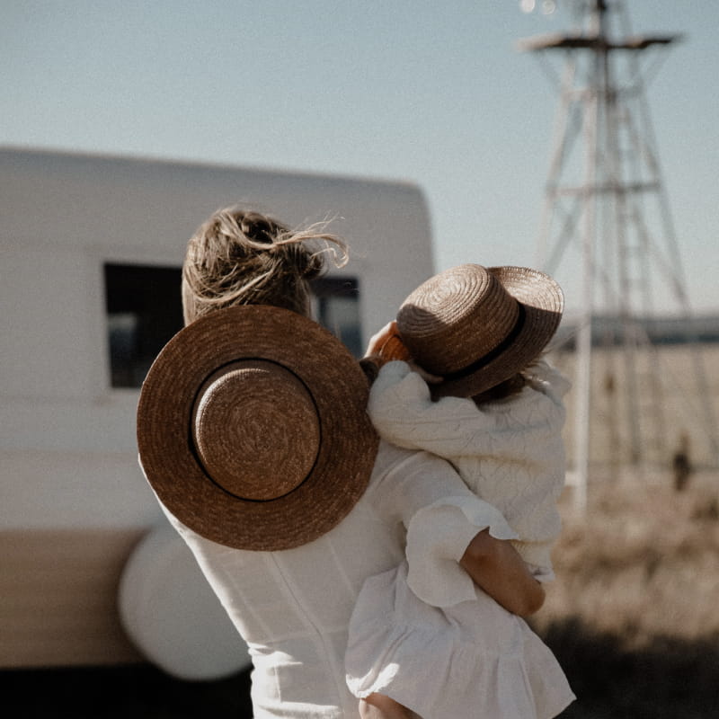 A close up of the back of a mother holding her child, both are wearing straw hats