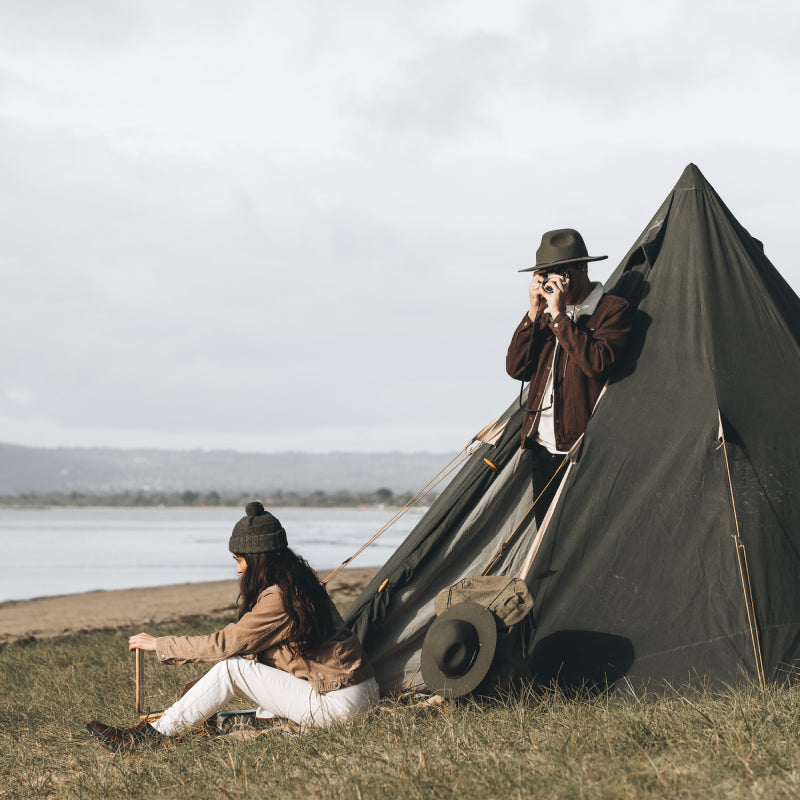 A man and a woman outside a bell tent on the beach, the man is taking a photo with his film camera