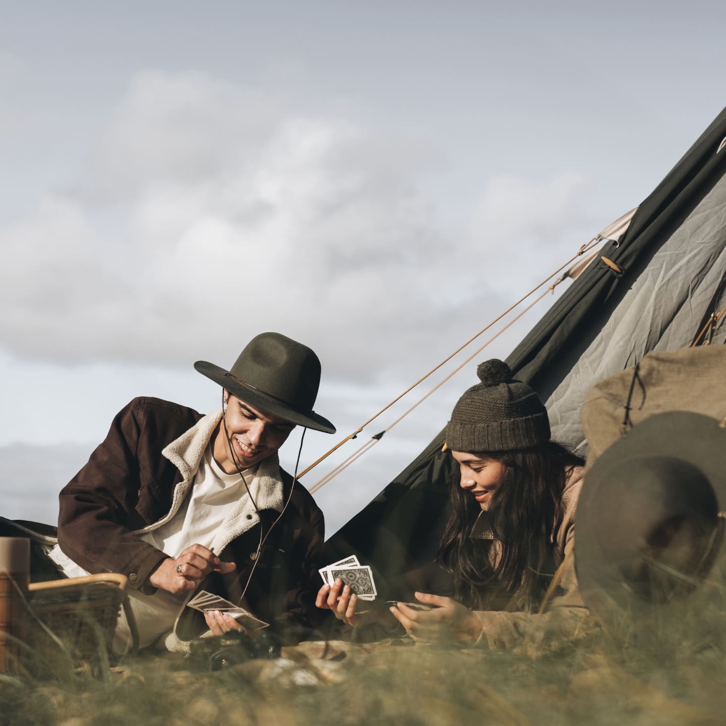 A man and a woman sitting in front of a tent playing cards on a picnic blanket