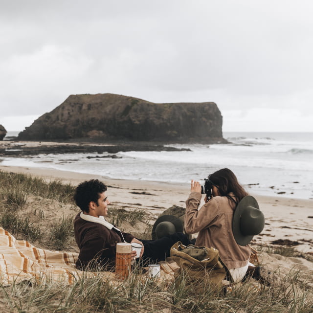 A man and a woman having a picnic on the beach, the woman is taking a photo of the man